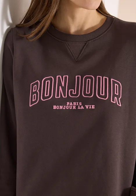 Frau trägt dunkelbraunes Sweatshirt mit Aufdruck: BONJOUR PARIS BONJOUR LA VIE.